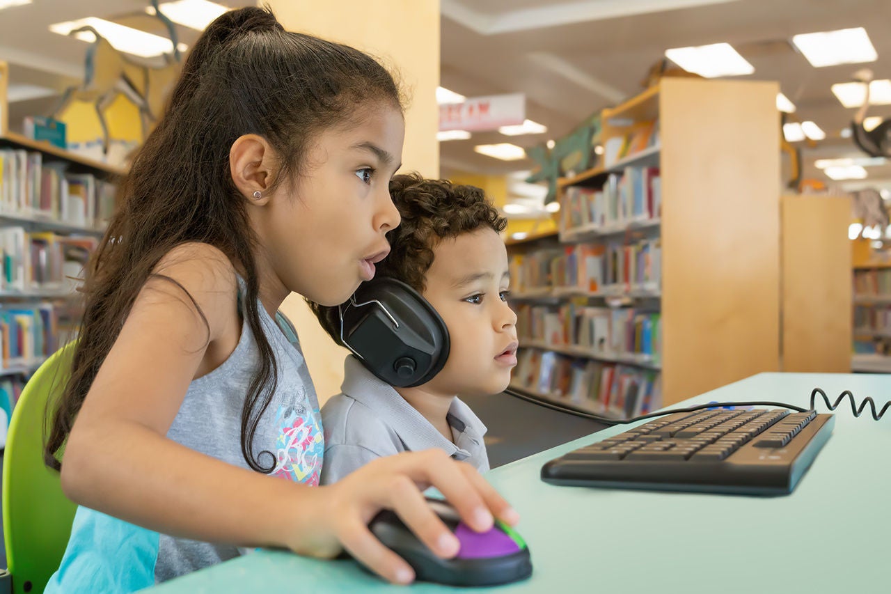 He wears the adult size headphones while she navigates with a wireless mouse in front of the computer screen.