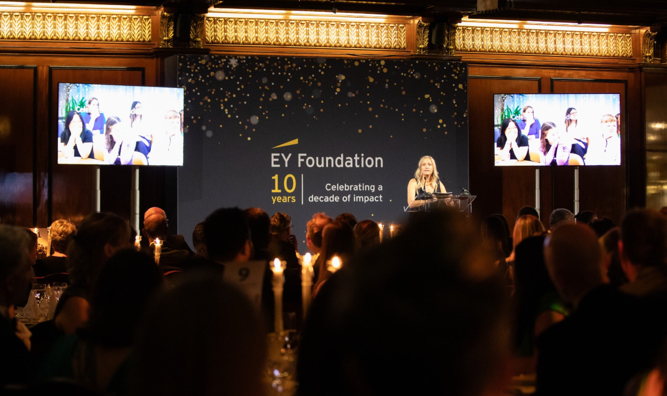 Lynne Peabody speaks on stage at a celebration dinner with the back of audiences heads in the bottom of the shot.