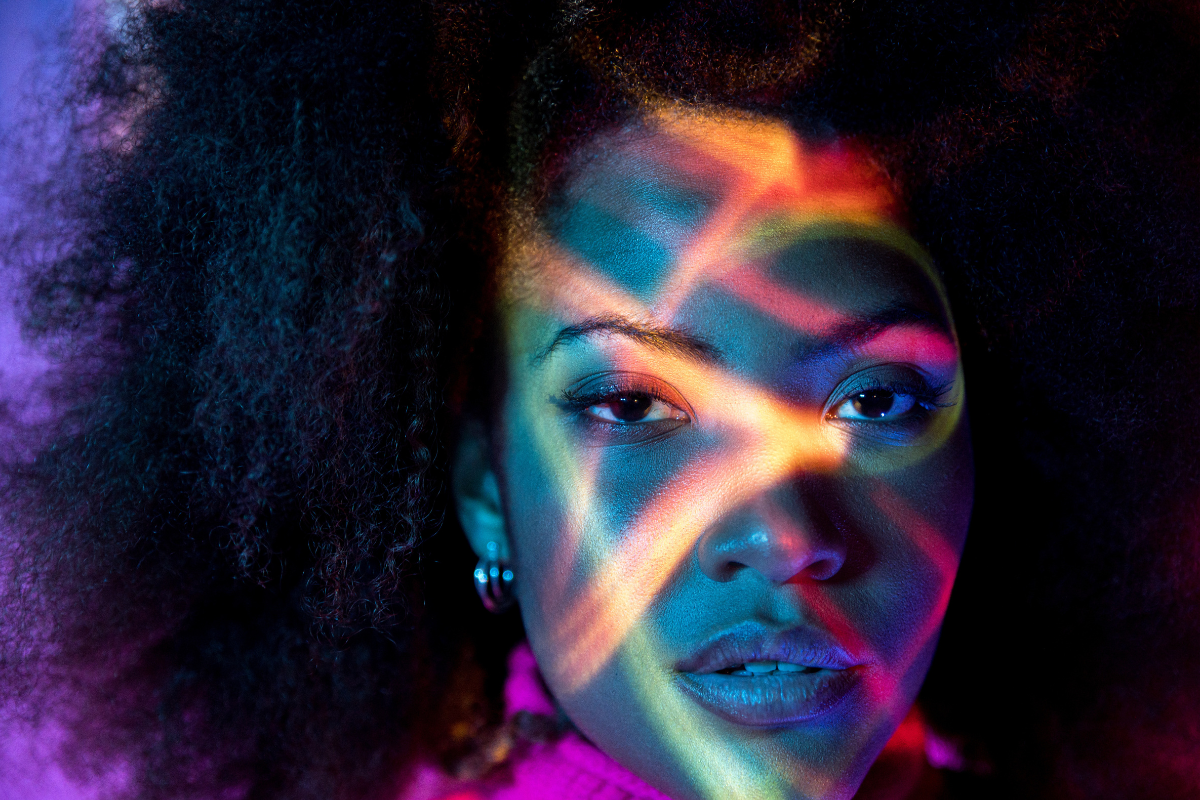 Image of a woman's face with a pink jumper and colourful lights like the reflections of a wave across her face,